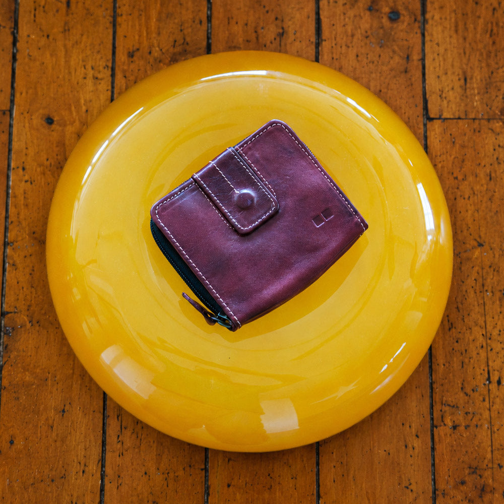 Burgundy leather bifold wallet with contrast stitching, snap tab, and zip compartment, resting on a glossy yellow round surface.