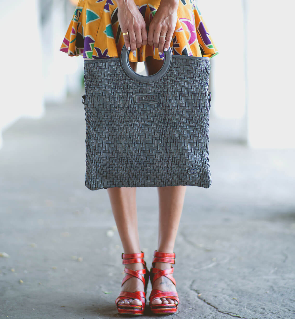 A woman wearing a colorful dress and sandals holding a Bed Stu Adele woven tote.