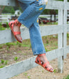 A woman wearing Afrodita jeans and Bed Stu sandals leaning against a fence.