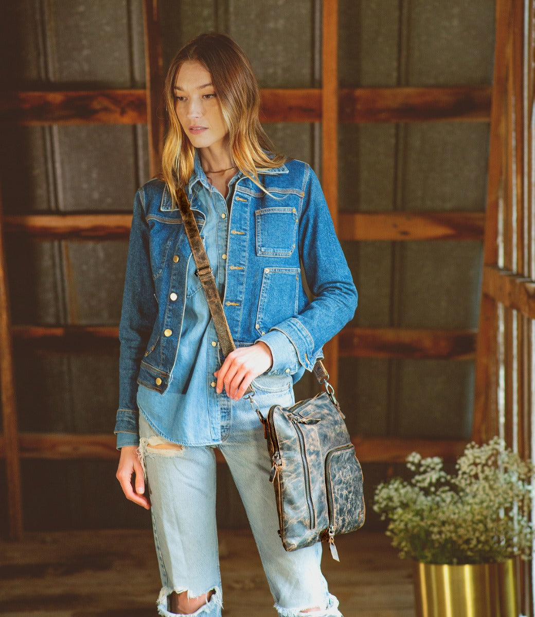 A woman wearing jean jacket, blue jeans and distressed black Aiken bag while standing in a barn.
