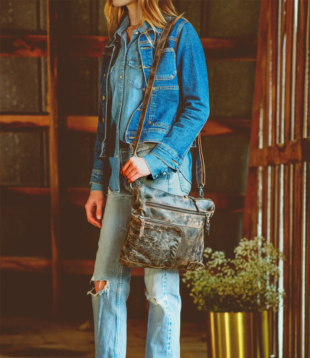 A woman wearing jean jacket, blue jeans and distressed black Aiken bag while standing in a barn.