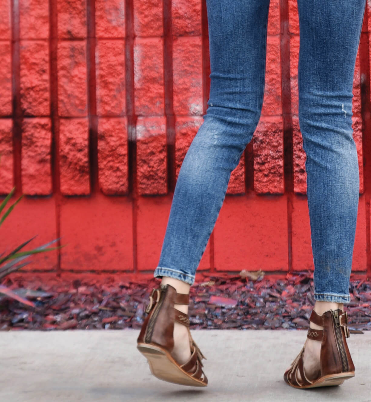 A woman wearing Alena jeans and Bed Stu sandals standing in front of a red brick wall.
