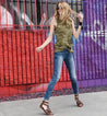 A woman wearing an Alena tank top from Bed Stu, jeans, and sandals in front of a graffiti wall.