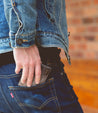A man in a denim jacket is holding a Bed Stu wallet in his pocket.