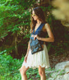 A woman in a white skirt and black Andie purse standing in the woods.
