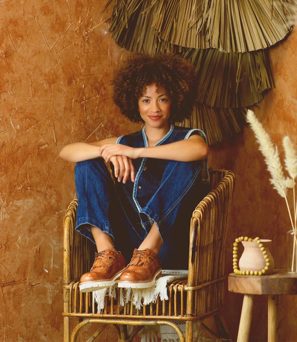 A woman sitting in a wicker chair while wearing blue jeans and tan Anise shoes..
