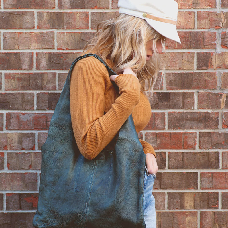 A woman wearing a hat and jeans holding a green leather Bed Stu Ariel tote bag.