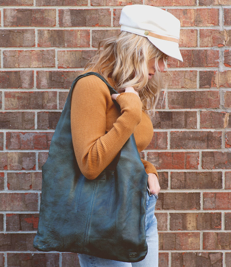 A woman wearing a hat and jeans holding a green leather Bed Stu Ariel tote bag.