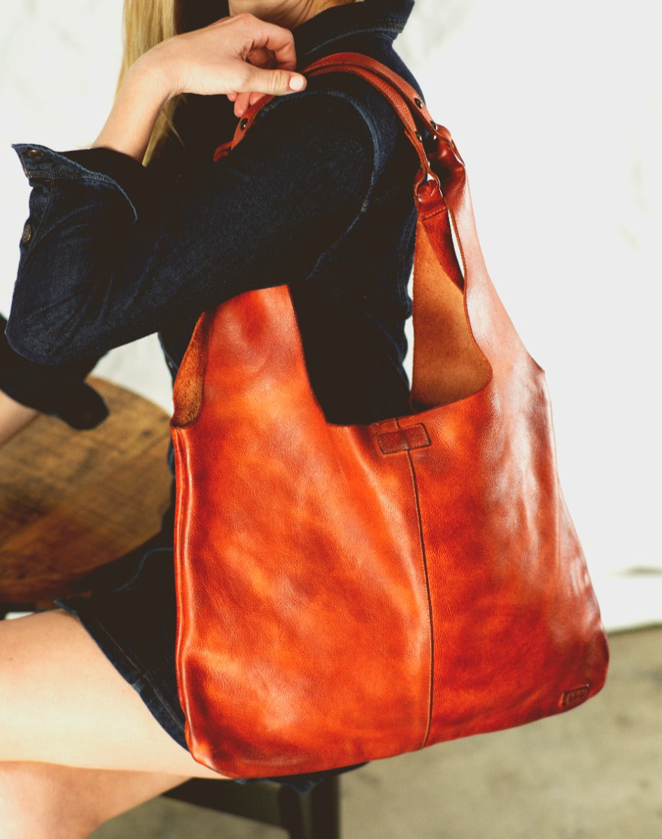 A woman is sitting on a chair holding a Bed Stu Ariel red leather bag.