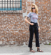 A woman is standing in front of a brick wall wearing Bed Stu jeans and a Bed Stu t-shirt.