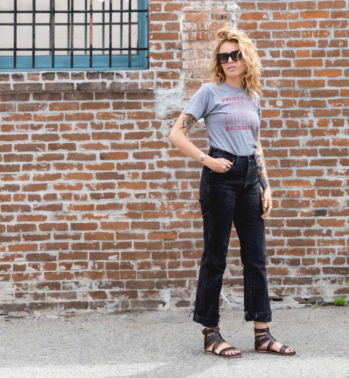 A woman is standing in front of a brick wall wearing Bed Stu jeans and a Bed Stu t-shirt.