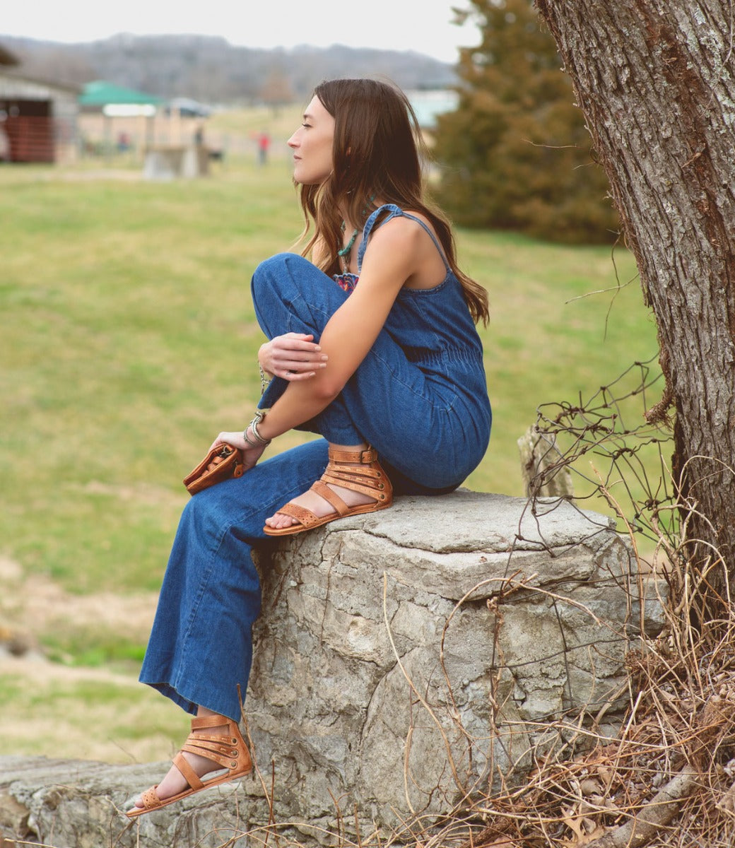 A woman sitting on top of a tree in a Bed Stu Artemis denim overall jumpsuit.