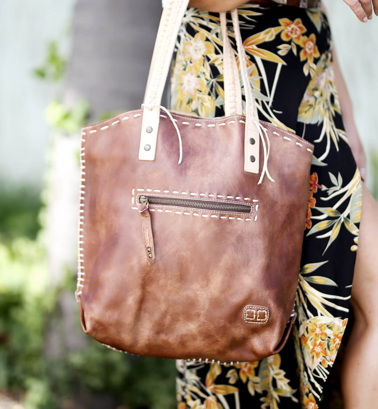 A close-up of a brown Bed Stu Barra handmade leather tote bag with contrast stitch detail and a zipper pocket, held by a person wearing a floral dress.