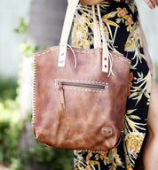 A close-up of a brown Bed Stu Barra handmade leather tote bag with contrast stitch detail and a zipper pocket, held by a person wearing a floral dress.