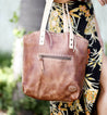A close-up of a brown Bed Stu Barra handmade leather tote bag with contrast stitch detail and a zipper pocket, held by a person wearing a floral dress.
