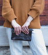 A woman wearing a tan sweater and white pants holding a Bed Stu Bayshore clutch.