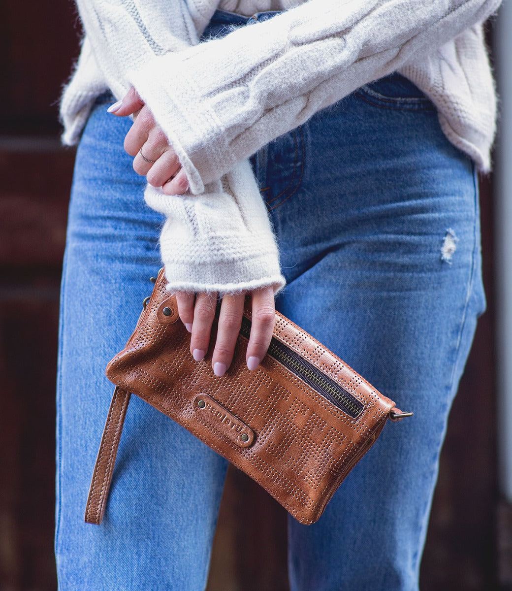 A woman wearing jeans and a sweater holding a Bed Stu Bayshore brown leather clutch.