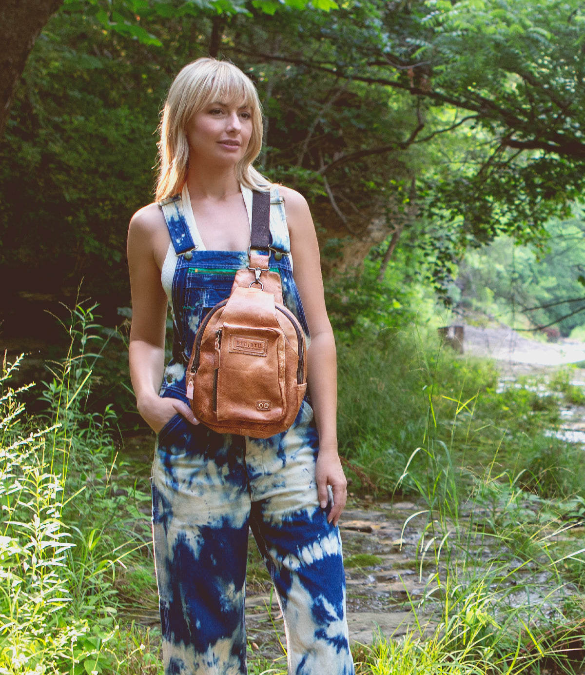 A woman in overalls standing in a wooded area with a Bed Stu Beau backpack.