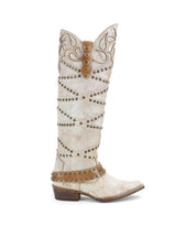 The Bed Stu "Betting" is a tall, white Western-style boot crafted from vegetable-tanned leather, decorated with brown harness straps and studs. It includes a patterned upper and a low heel, offering rustic elegance to any outfit.