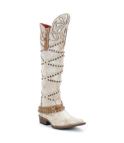 The Bed Stu Betting is a tall white Western boot made of vegetable-tanned leather, featuring decorative stitching, studs, a flower pattern near the top, a low heel, and an elegant harness strap for added style.