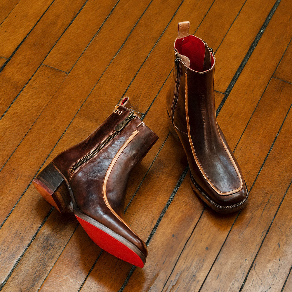 Pair of dark brown leather ankle boots with tan piping, side zippers, and red soles arranged on a wooden floor.