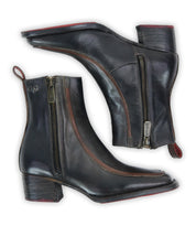 The Bed Stu Bitsy is a pair of black hand-finished leather ankle boots featuring side zippers, brown trim accents, stacked wooden heels, and red pull tabs, shown on a white background.