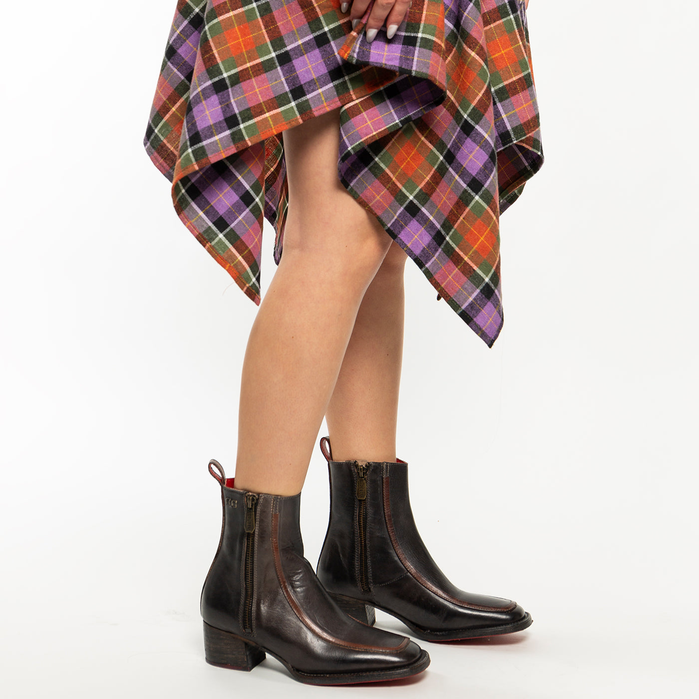 A person wearing Bed Stu's Bitsy dark brown ankle boots with a stacked wooden heel and a colorful plaid skirt stands against a plain white background.