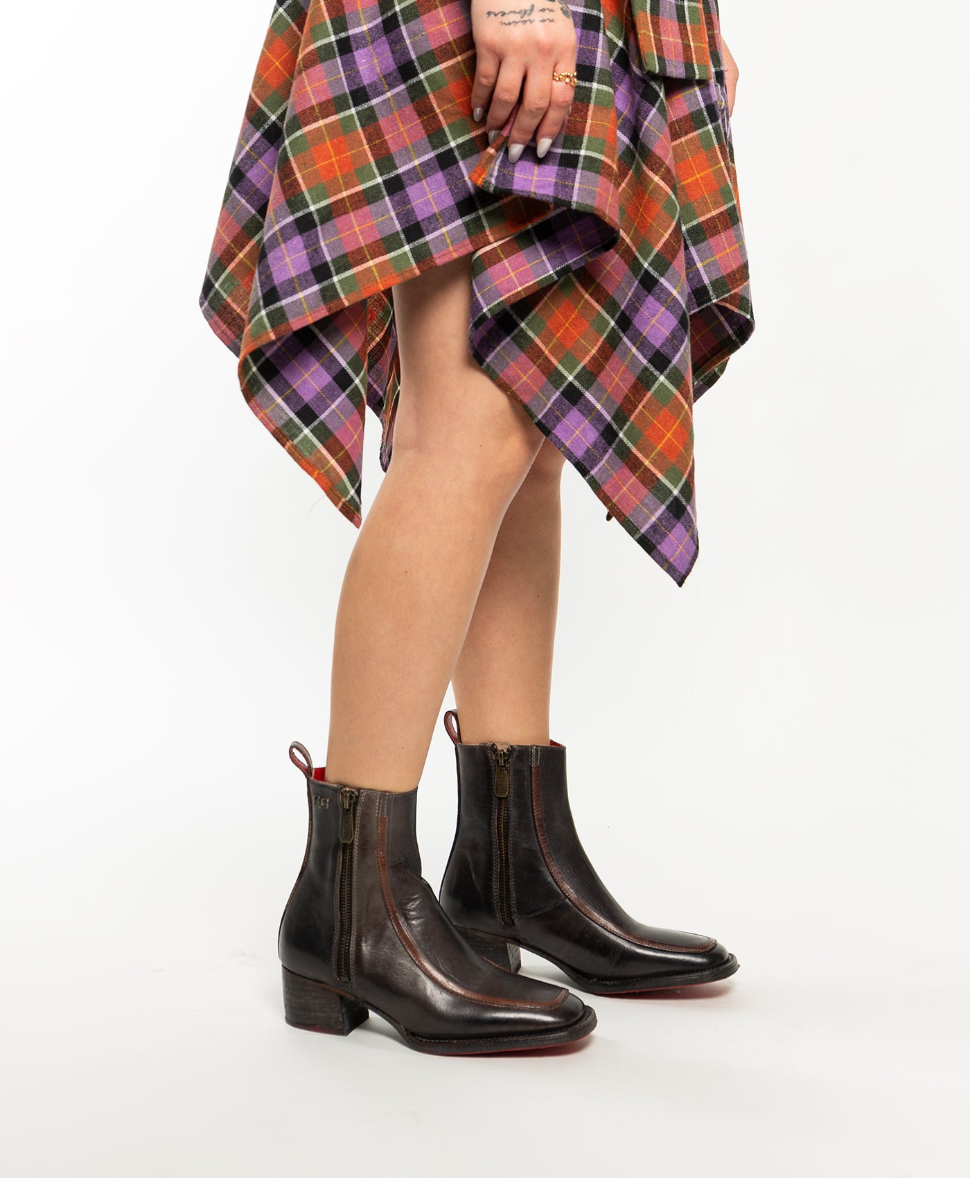 A person wearing Bed Stu's Bitsy dark brown ankle boots with a stacked wooden heel and a colorful plaid skirt stands against a plain white background.