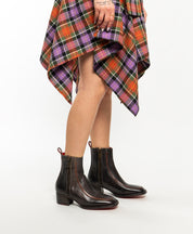 A person wearing Bed Stu's Bitsy dark brown ankle boots with a stacked wooden heel and a colorful plaid skirt stands against a plain white background.