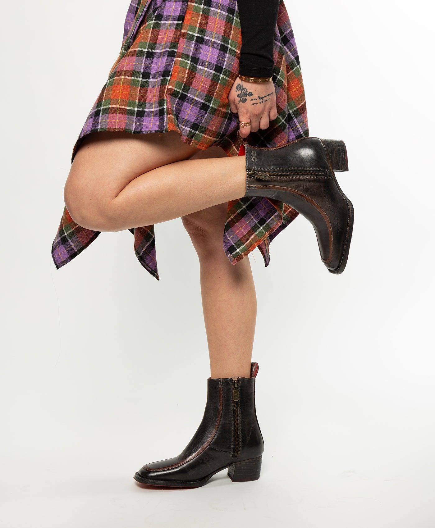 A person stands on one leg, holding their other ankle, wearing a plaid skirt and Bed Stu Bitsy black hand-finished leather ankle boots with a stacked wooden heel. Tattoos are visible on their hand.