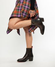 A person stands on one leg, holding their other ankle, wearing a plaid skirt and Bed Stu Bitsy black hand-finished leather ankle boots with a stacked wooden heel. Tattoos are visible on their hand.