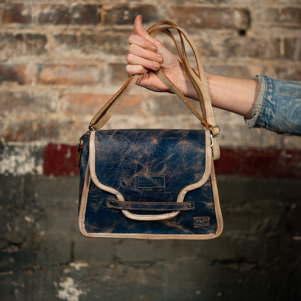 Hand holding a distressed navy leather crossbody bag with cream trim and straps against a weathered brick wall.