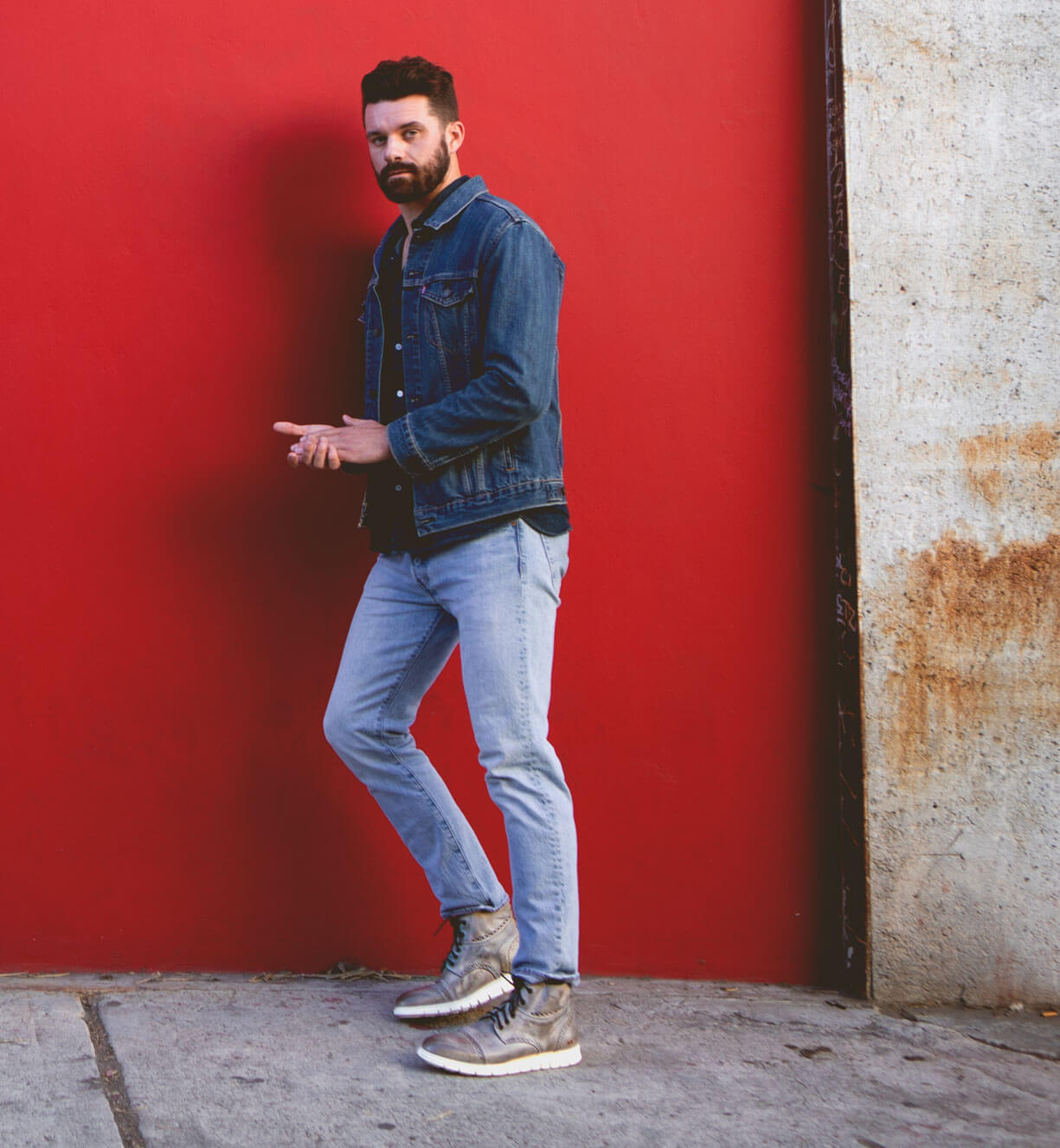 A man in a Bed Stu denim jacket leaning against a red wall.