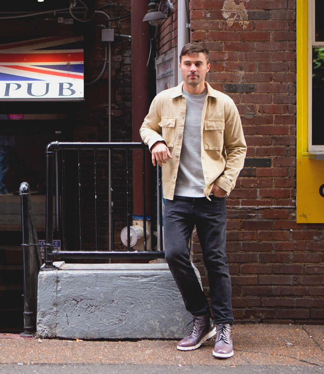 A man in a tan Bowery II jacket leaning against a brick wall, made by Bed Stu.