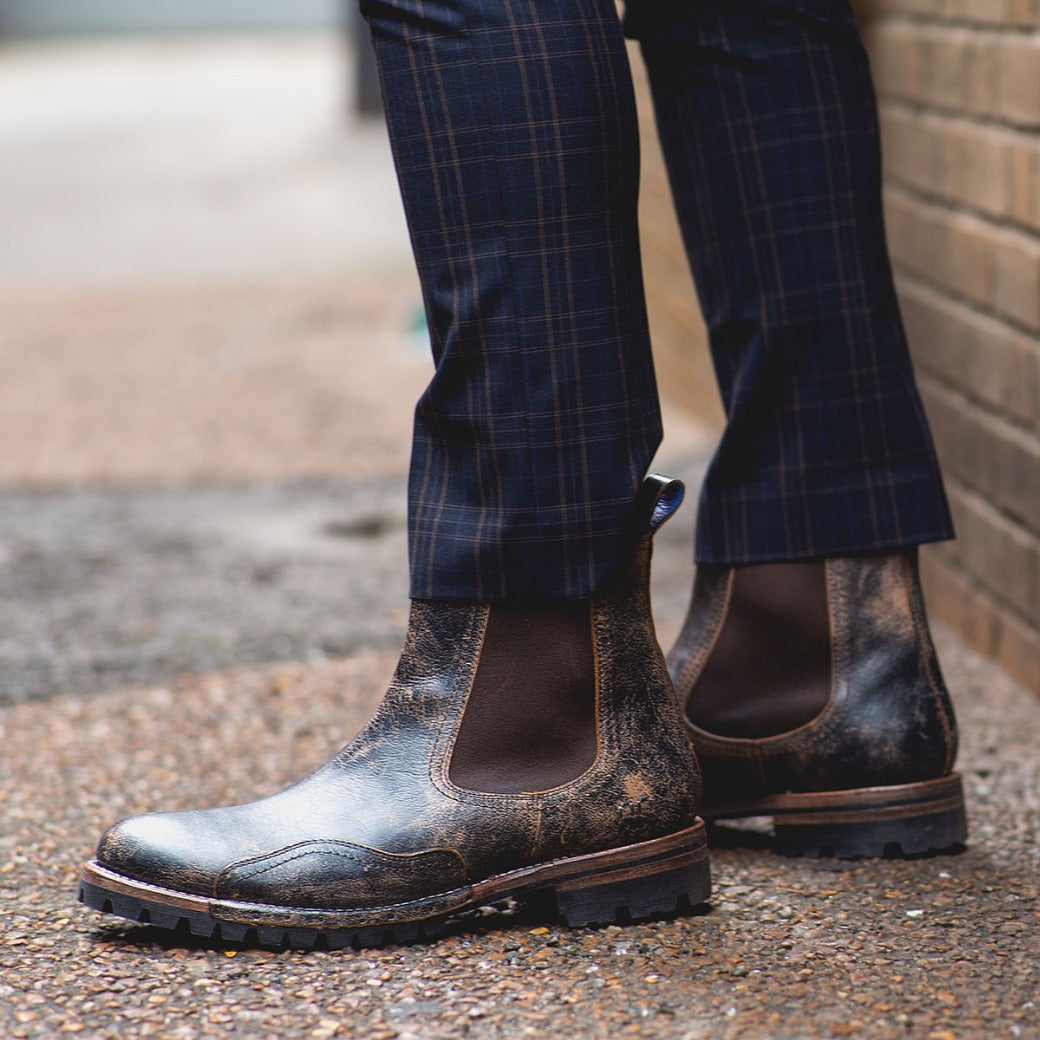 A person wearing Bed Stu Brady Trek boots with a Vibram outsole and plaid trousers standing on a brick-lined sidewalk.