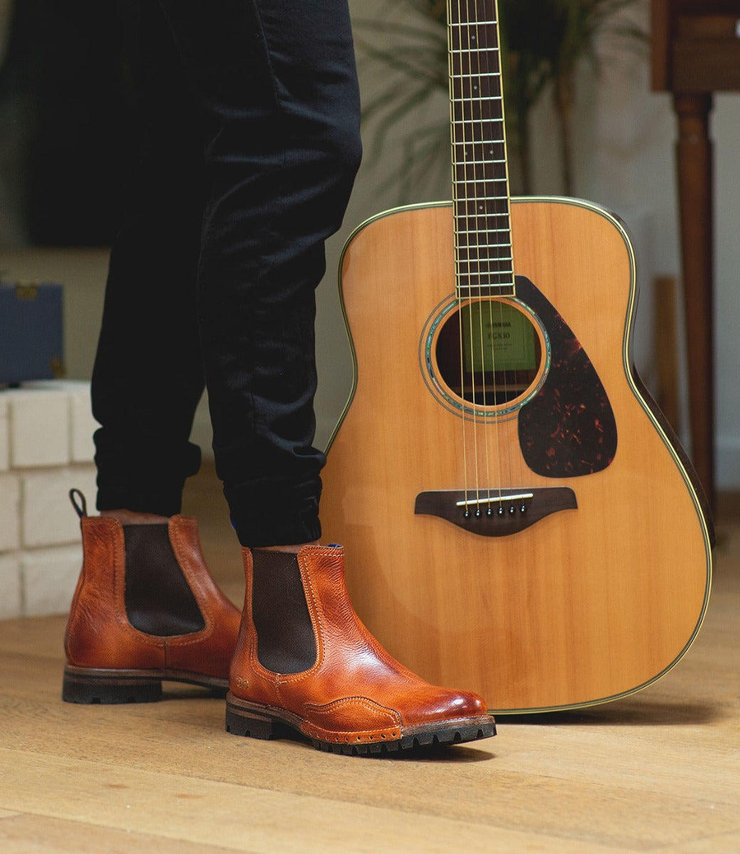 A man wearing a Brady Trek boots by Bed Stu standing next to an acoustic guitar.