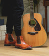 A person standing next to an Brady Trek acoustic guitar, wearing shiny brown Bed Stu chelsea boots with a Vibram outsole on a wooden floor.