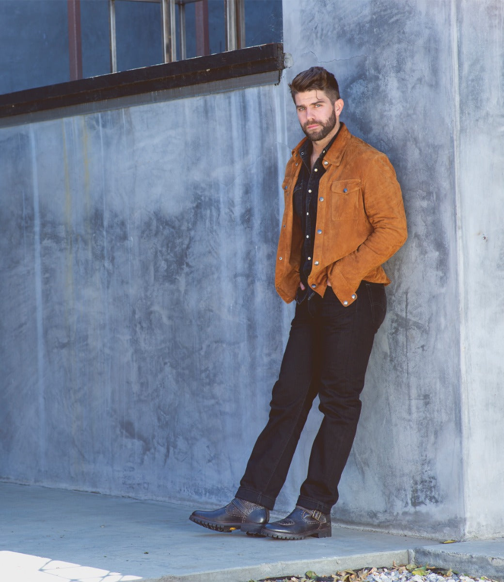 A man in a brown Bed Stu jacket leaning against a wall.