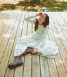 A woman in a white dress sitting on a wooden deck wearing Brianna by Bed Stu.