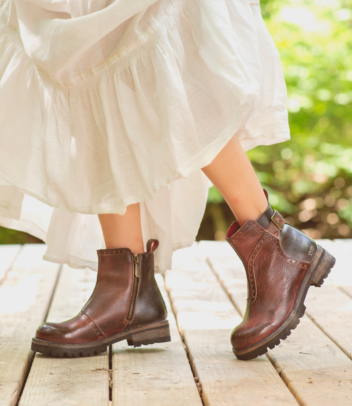 A woman in a white dress is standing on a wooden deck, wearing  Brianna boots by Bed Stu.