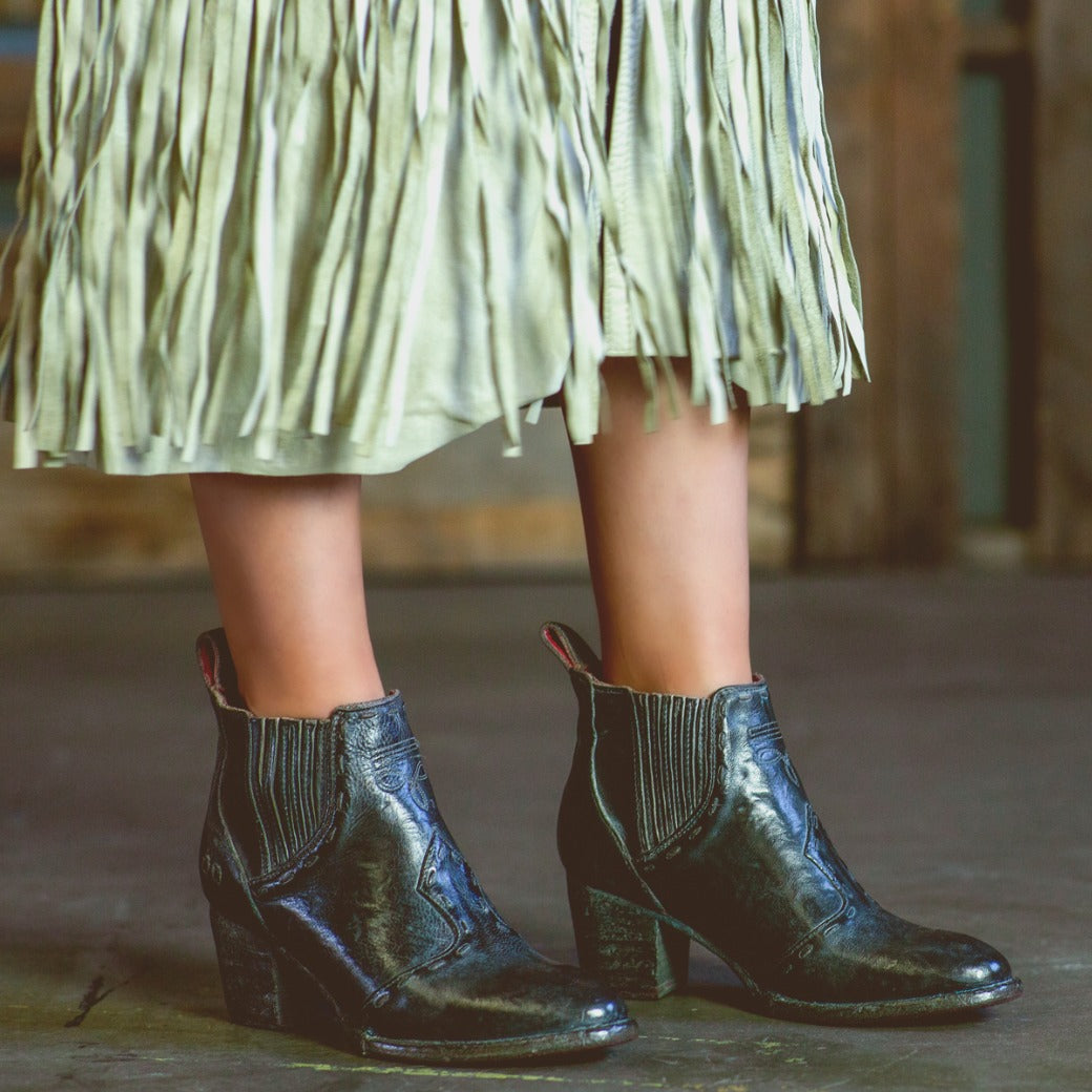 A woman wearing black Bed Stu Brie II boots and a fringe skirt.