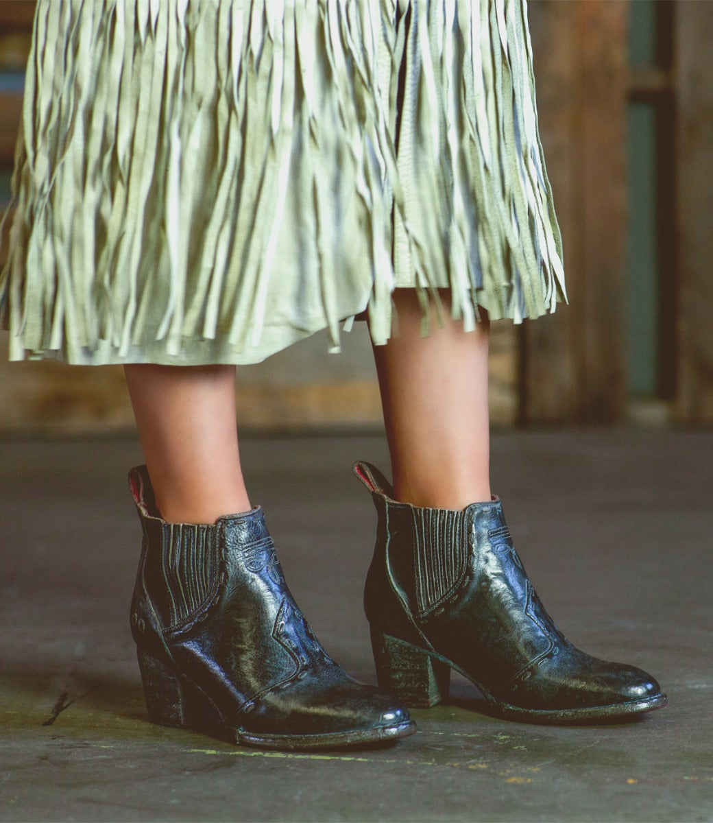 A woman wearing black Bed Stu Brie II boots and a fringe skirt.