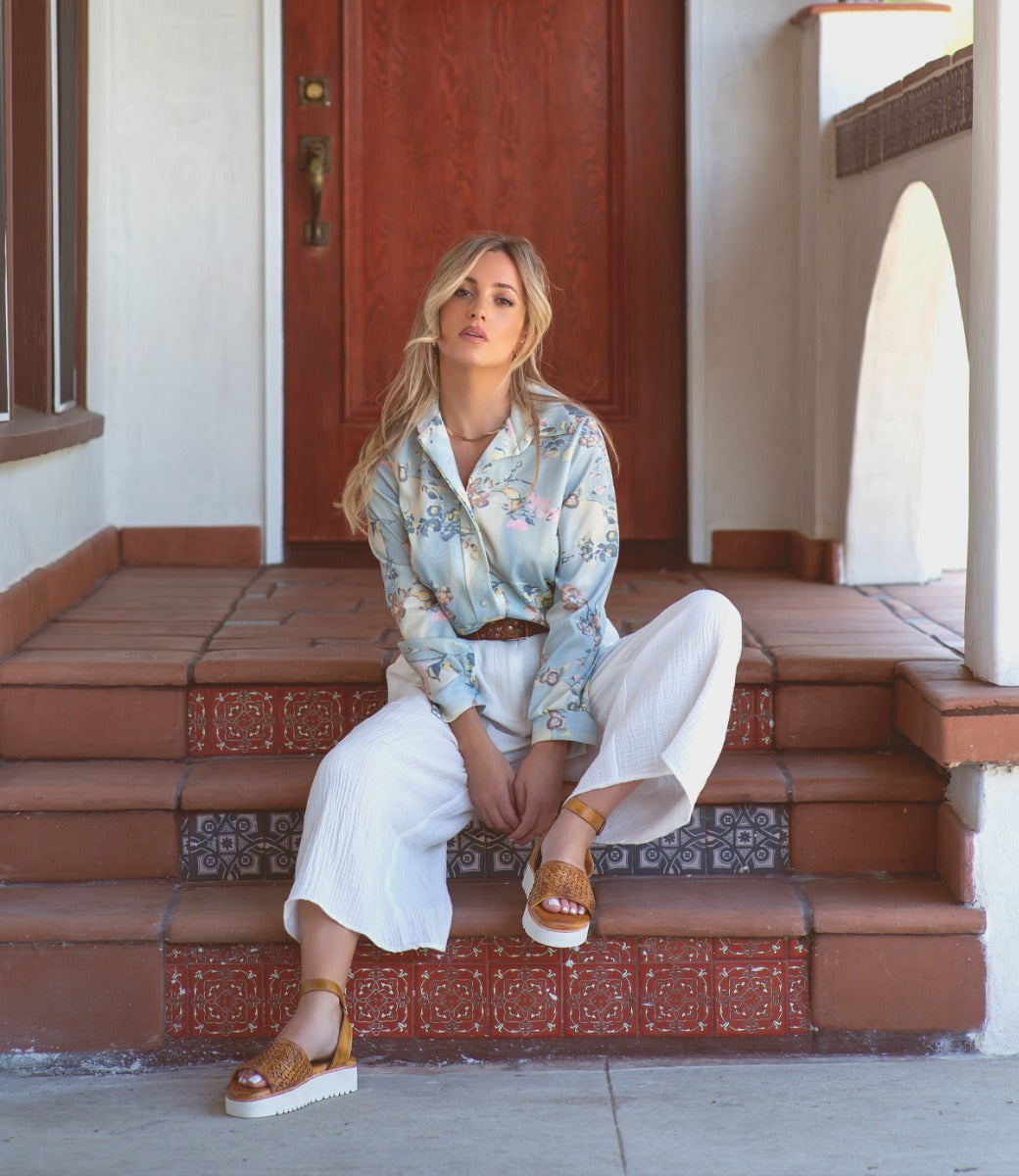 A woman sitting on the steps of a house wearing a Brisa floral shirt and Bed Stu sandals.