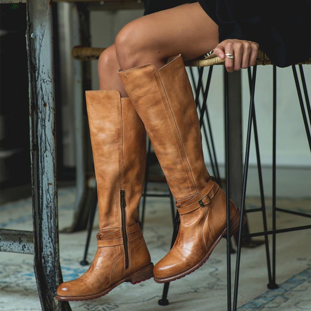 A woman sitting on a stool wearing Bed Stu tan leather boots.