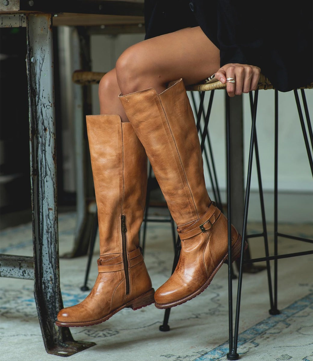 A woman sitting on a stool wearing Bed Stu tan leather boots.