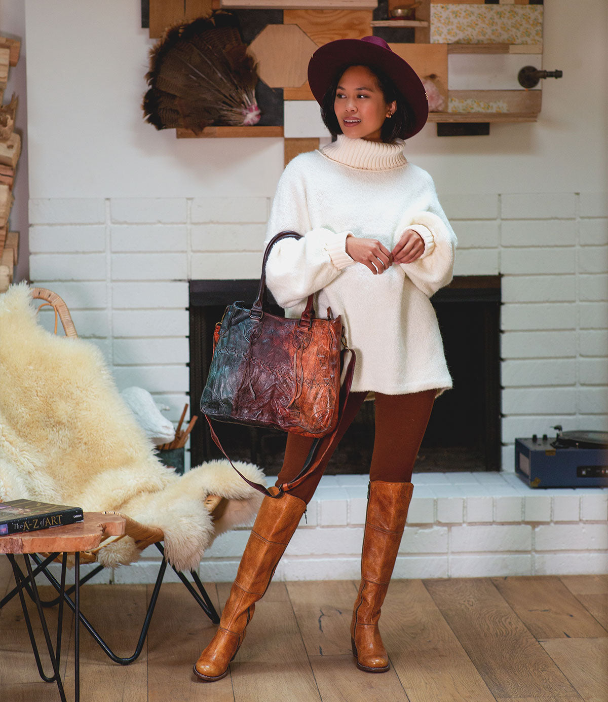 A person in a white turtleneck, brown leggings, and tall boots stands indoors holding the Bed Stu Bruna, a large artisanal leather bag, and wearing a wide-brimmed hat.