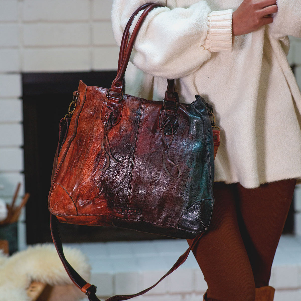 A person in a cream sweater and brown leggings holds the Bed Stu Bruna, a large two-toned leather bag with double handles and shoulder strap, highlighting its artisanal craftsmanship.