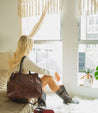 A woman sitting on a couch with a brown Bed Stu Bruna leather bag.