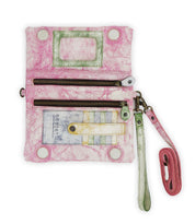 The Cadence by Bed Stu is a compact wallet with a pink and white pattern. It features two zippered compartments, a transparent ID slot, and an attached strap. Its versatile 3-in-1 design easily converts it into a crossbody for added convenience.