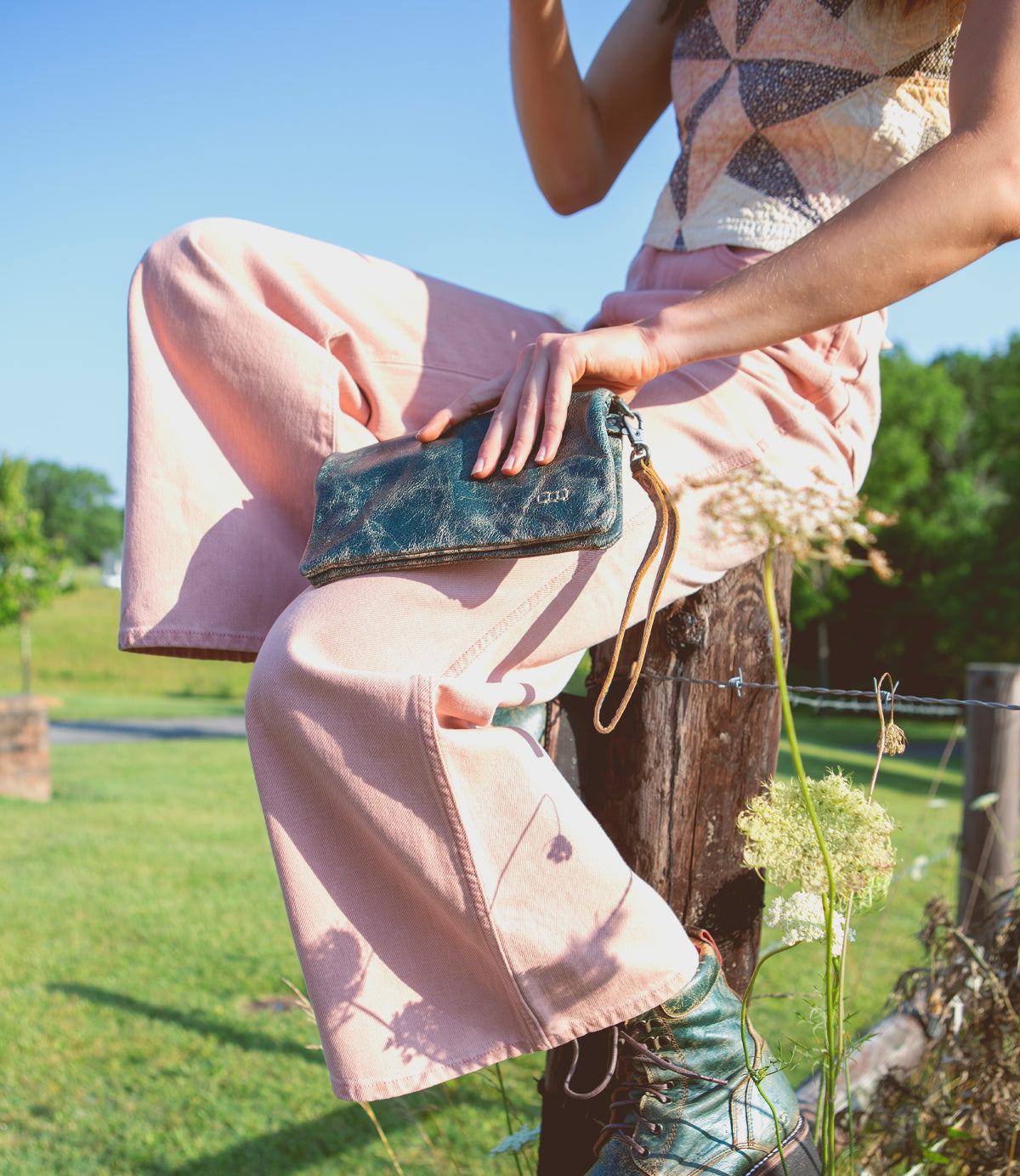 A woman is sitting on a wooden post holding a Bed Stu Cadence clutch.